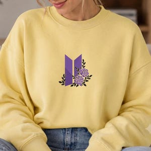 Può includere: Una felpa girocollo giallo pallido con un ricamo centrale raffigurante un logo geometrico viola affiancato da due rose viola con contorni neri e foglie nere. Il capo presenta maniche lunghe e bordi a coste su colletto, polsini e orlo.