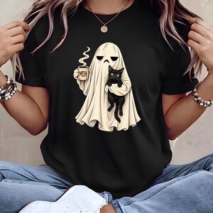 May include: A black t-shirt featuring a graphic of a white ghost holding a steaming mug with the text MEH printed on it. The ghost is also holding a small black cat. The design is centered on the chest of the shirt, showcasing a whimsical illustration.