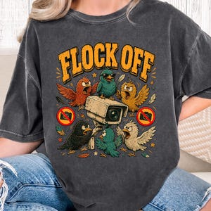 Peut inclure: Un t-shirt gris anthracite à manches courtes avec une impression graphique représentant plusieurs oiseaux colorés entourant une caméra de sécurité. Le texte FLOCK OFF est imprimé en grandes lettres jaunes au-dessus, avec deux symboles d'interdiction.