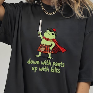 May include: A black t-shirt featuring a graphic of a green frog dressed in a red plaid kilt, a matching cape, and a tam o' shanter hat, holding a sword. Below the frog, the text reads, "down with pants up with kilts" in light green letters.