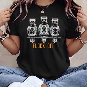 以下が含まれることがあります： ワイヤーにとまった3羽のロボット鳥のグラフィックが描かれた黒いTシャツ。各鳥の目はカメラレンズ、頭部はスクリーンになっています。鳥の下にはオレンジ色でFLOCK OFFとプリントされており、各鳥にはGOVERNMENT PROPERTYという小さなラベルが付いています。