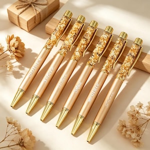 May include: Six gold-toned ballpoint pens arranged in a row. Each pen features a clear upper barrel filled with gold leaf flakes, a metallic gold lower barrel with the word Solace printed in white, and a gold clip and tip.