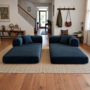 May include: Two matching dark blue corduroy chaise lounges positioned side by side on a textured beige area rug. Each piece features a low-profile rectangular base, a single rounded armrest on the outer side, and one square throw pillow.