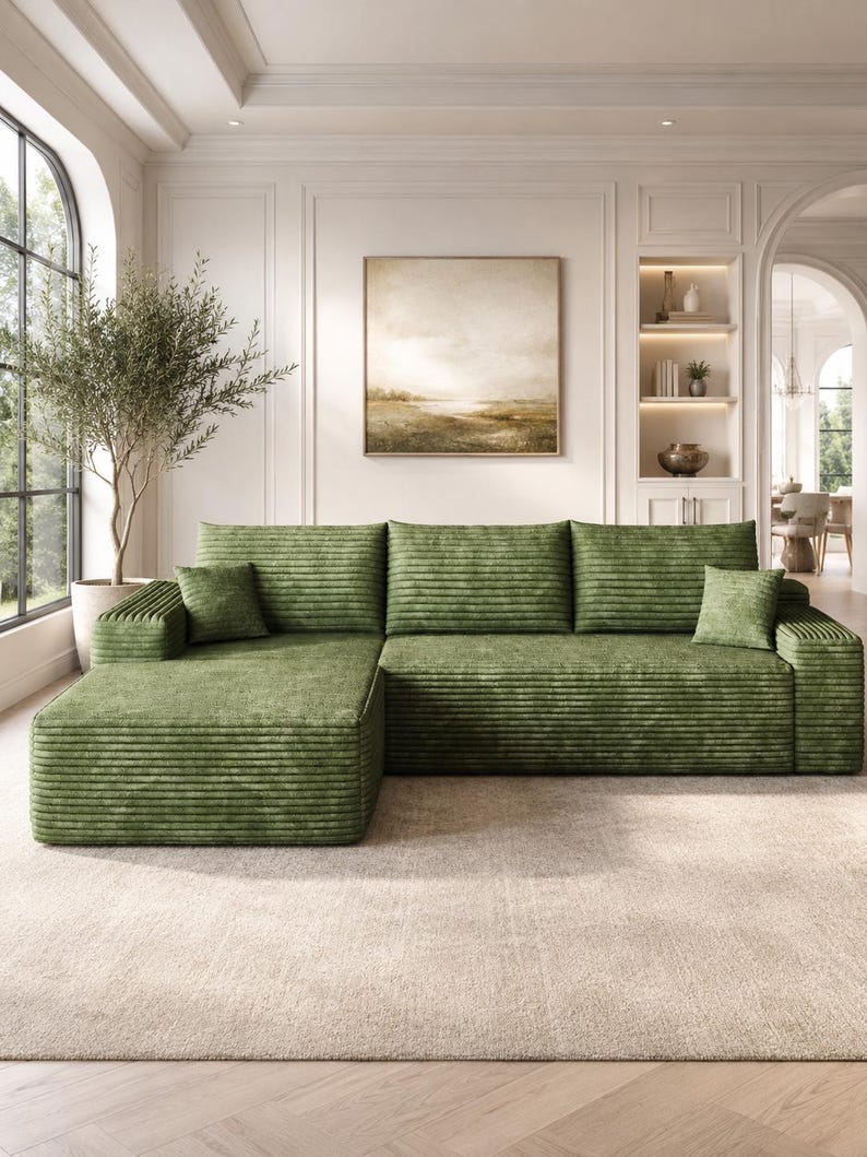 May include: A large, L-shaped sectional sofa upholstered in a textured, olive green corduroy fabric. The sofa features a chaise lounge on the left side, three back cushions, and two small square throw pillows in the same matching green fabric.