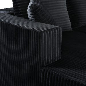 Op de afbeelding: Een zwarte bank met een dikke, verticaal geribbelde corduroy textuur. Het meubelstuk is voorzien van een prominente vierkante armleuning en bijpassende kussens van hetzelfde geribbelde materiaal, met een diepe, uniforme donkere kleur over de gehele structuur.