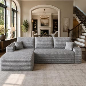May include: A large light gray sectional sofa featuring a horizontal ribbed texture across the cushions and frame. The sofa includes a chaise lounge on the left side and two matching square throw pillows, providing a structured seating arrangement.