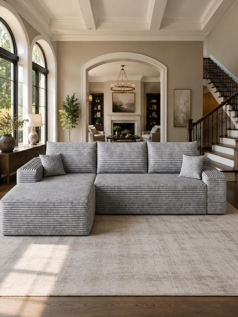 May include: A large light gray sectional sofa featuring a horizontal ribbed texture across the cushions and frame. The sofa includes a chaise lounge on the left side and two matching square throw pillows, providing a structured seating arrangement.