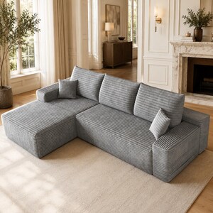 May include: A light gray sectional sofa featuring a textured corduroy fabric, a chaise lounge section on the left, three large back cushions, and two small square throw pillows. The sofa has a low-profile design with wide, flat armrests.