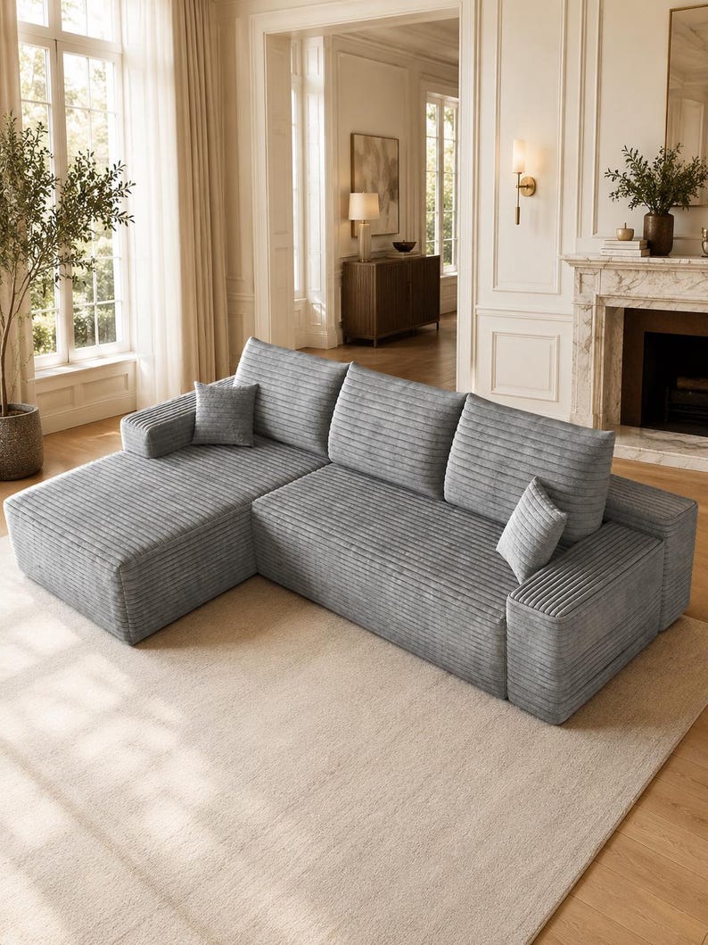 May include: A light gray sectional sofa featuring a textured corduroy fabric, a chaise lounge section on the left, three large back cushions, and two small square throw pillows. The sofa has a low-profile design with wide, flat armrests.