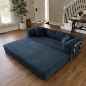 May include: A deep blue corduroy sofa bed featuring a wide, low-profile base and a backrest with two matching square throw pillows. The furniture piece has a textured ribbed fabric finish and is supported by a sleek, silver-toned metal frame.