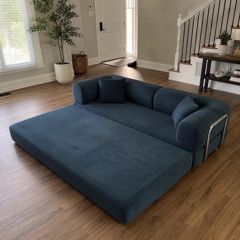 May include: A deep blue corduroy sofa bed featuring a wide, low-profile base and a backrest with two matching square throw pillows. The furniture piece has a textured ribbed fabric finish and is supported by a sleek, silver-toned metal frame.