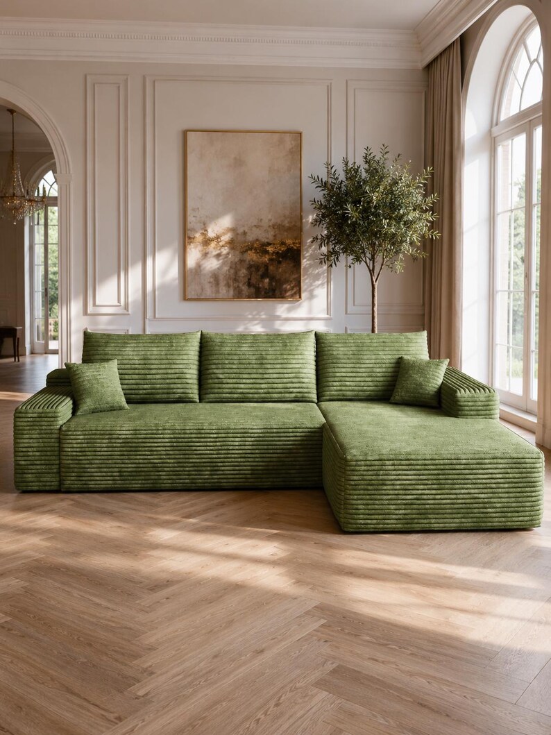 May include: A large, L-shaped sectional sofa upholstered in a textured, olive green corduroy fabric. The sofa features a long chaise lounge on the right side, three wide back cushions, and two small, square throw pillows in the same green fabric.