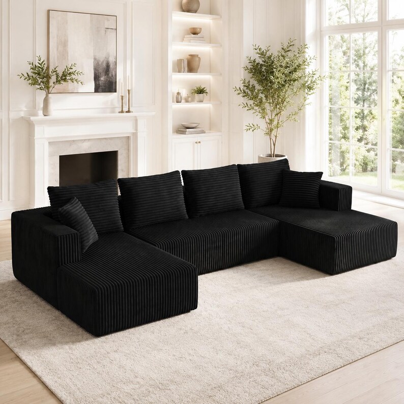 Op de afbeelding: Een grote zwarte hoekbank van gestructureerde corduroy stof. De bank heeft een U-vormige configuratie met twee verlengde chaise longue-gedeelten, diepe zittingen, meerdere bijpassende vierkante rugkussens en &eacute;&eacute;n klein sierkussen.