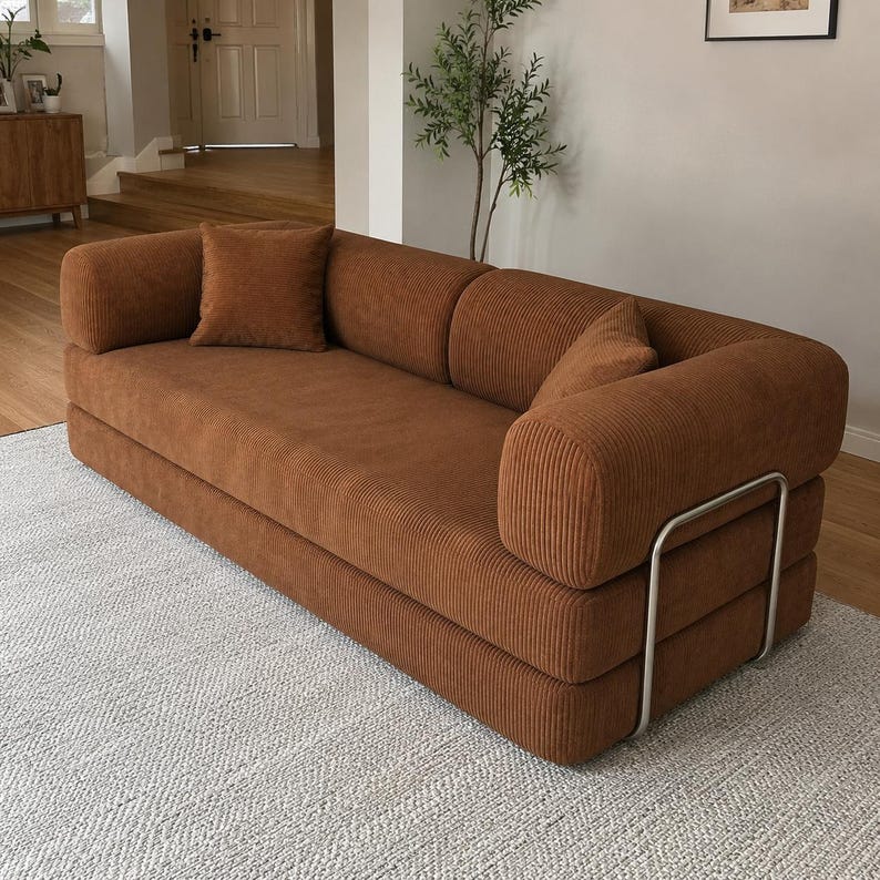 May include: A three-seater sofa upholstered in brown corduroy fabric. The sofa features rounded armrests, a low backrest, and two matching square throw pillows. A silver-toned metal frame supports the exterior sides of the sofa structure.