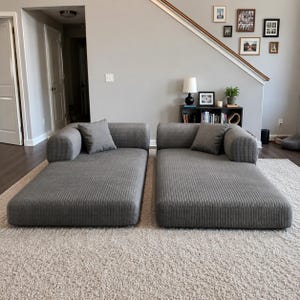 May include: Two matching gray corduroy chaise lounges, each featuring a low-profile rectangular base, a single rounded armrest on the outer side, and one square throw pillow in a coordinating gray fabric, arranged side by side on a light rug.