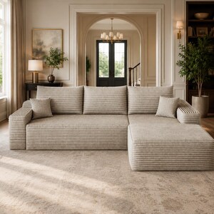May include: A large beige sectional sofa with a textured, horizontally ribbed fabric finish. The sofa features three back cushions, a chaise lounge section on the right side, and two small square throw pillows placed at the ends of the seating area.