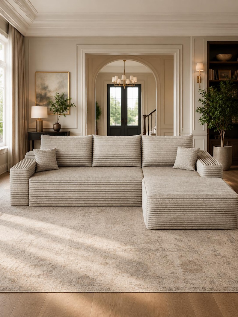 May include: A large beige sectional sofa with a textured, horizontally ribbed fabric finish. The sofa features three back cushions, a chaise lounge section on the right side, and two small square throw pillows placed at the ends of the seating area.