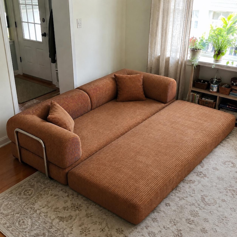 May include: A rust-colored corduroy sofa bed featuring a low-profile design with a pull-out section, two matching square throw pillows, and a silver-toned metal frame support on the side, providing a wide seating and sleeping surface area.