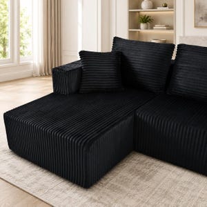 Op de afbeelding: Een zwarte hoekbank bekleed met een gestructureerde corduroy stof met verticale ribbels. Het meubelstuk heeft een laag ontwerp, een brede armleuning en verschillende grote, vierkante rugkussens die passen bij de diepzwarte kleur.
