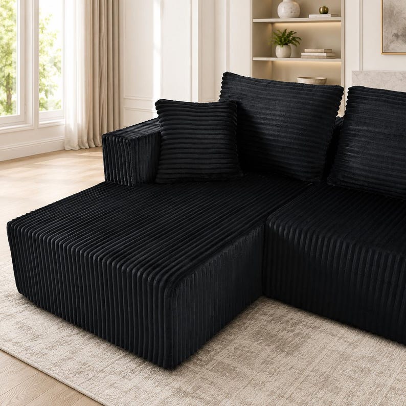 Op de afbeelding: Een zwarte hoekbank bekleed met een gestructureerde corduroy stof met verticale ribbels. Het meubelstuk heeft een laag ontwerp, een brede armleuning en verschillende grote, vierkante rugkussens die passen bij de diepzwarte kleur.