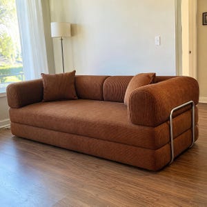 May include: A rust-colored corduroy sofa featuring a low-profile design with rounded armrests and a silver-toned metal frame support on the side. The sofa includes two matching square corduroy throw pillows placed against the back cushions.