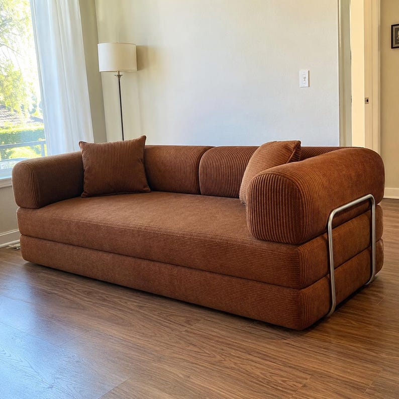 May include: A rust-colored corduroy sofa featuring a low-profile design with rounded armrests and a silver-toned metal frame support on the side. The sofa includes two matching square corduroy throw pillows placed against the back cushions.