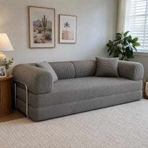 May include: A gray sofa with a ribbed texture on the rounded armrests and back cushions. The sofa features a low-profile design with a smooth seat and a silver metal frame visible on the side. Two matching square throw pillows are placed on the seat.