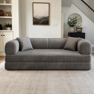 May include: A gray corduroy sofa featuring a low-profile design with rounded armrests and a smooth, continuous backrest. The piece includes two matching square corduroy throw pillows placed on either side of the seat, providing a uniform texture.