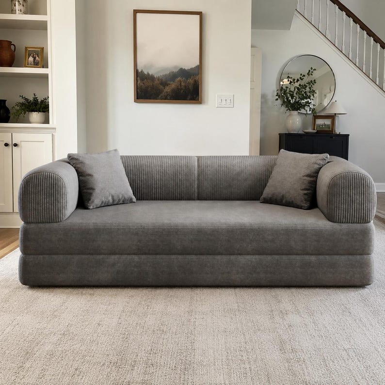 May include: A gray corduroy sofa featuring a low-profile design with rounded armrests and a smooth, continuous backrest. The piece includes two matching square corduroy throw pillows placed on either side of the seat, providing a uniform texture.