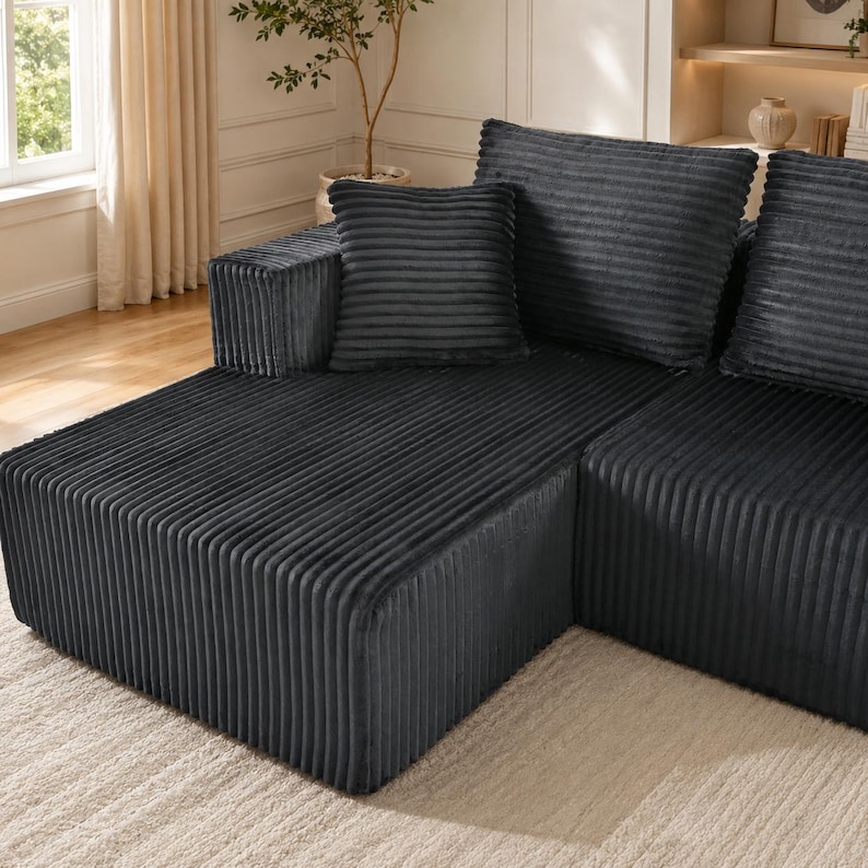 Op de afbeelding: Een donker antracietgrijze hoekbank van gestructureerde corduroy stof met verticale ribbeldetails. Het modulaire meubelstuk is voorzien van grote vierkante rugkussens en een lage basis, wat zorgt voor een strakke en uniforme uitstraling.
