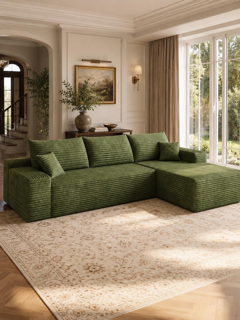 May include: A large, L-shaped sectional sofa upholstered in a textured, olive green corduroy fabric. The sofa features three wide back cushions, a chaise lounge section on the right side, and two small, square accent pillows in the same green fabric.