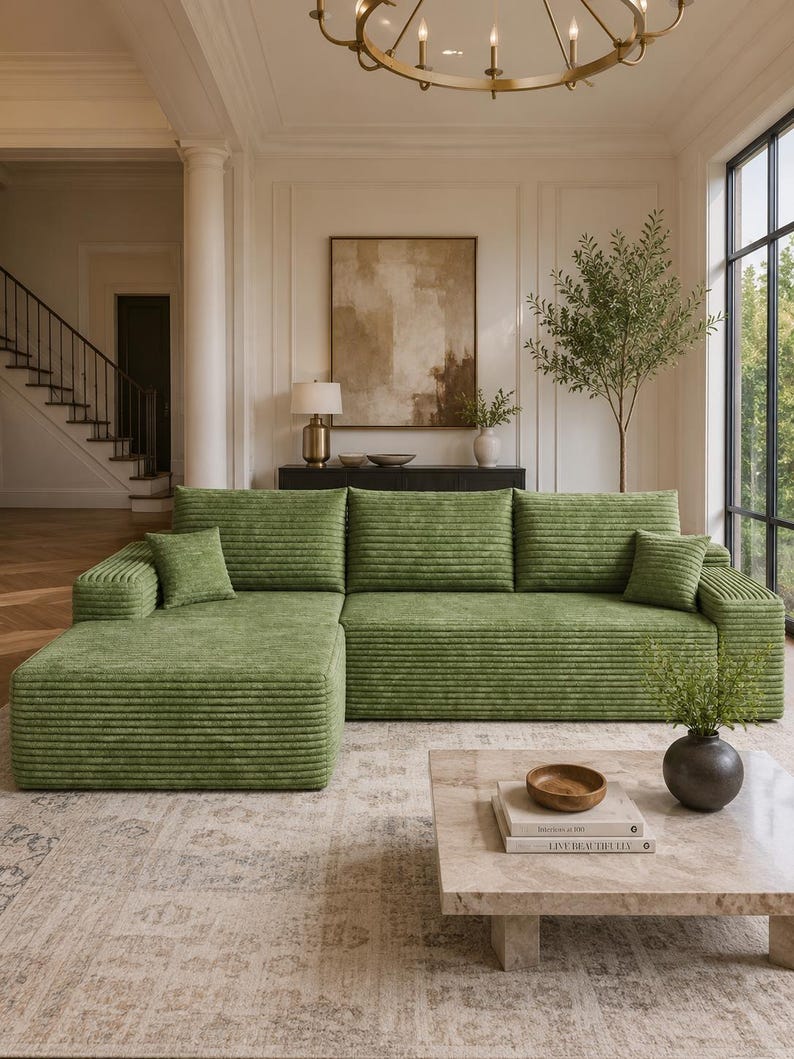 May include: A large, L-shaped sectional sofa upholstered in a textured, ribbed green fabric. The sofa features a chaise lounge on the left side, three back cushions, and two small square throw pillows placed at each end of the seating area.