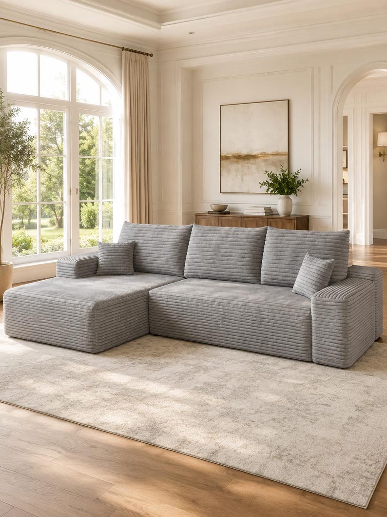 May include: A light gray sectional sofa featuring a textured corduroy fabric, a chaise lounge on the left side, three large back cushions, and two small square throw pillows. The sofa has a low-profile design with wide, flat armrests.