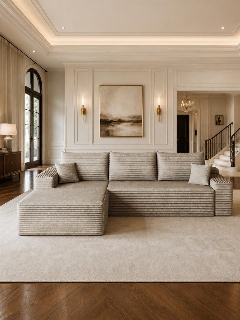 May include: A large, beige sectional sofa featuring a horizontal ribbed texture across the cushions and frame. The sofa includes a chaise lounge section on the left side and two small, matching square throw pillows placed on the seat cushions.