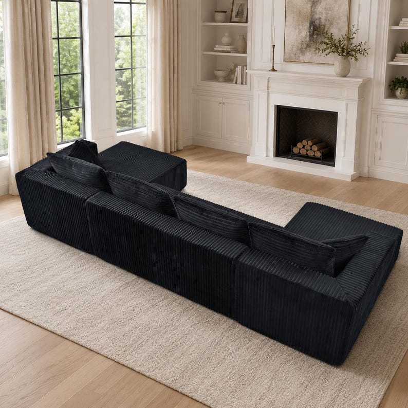 Op de afbeelding: Een grote, zwarte corduroy hoekbank met een laag profiel, voorzien van een modulaire configuratie met twee chaise longue-uiteinden, diepe zittingen en bijpassende rechthoekige rugkussens, met een textuurrijke, geribbelde stoffen afwerking.