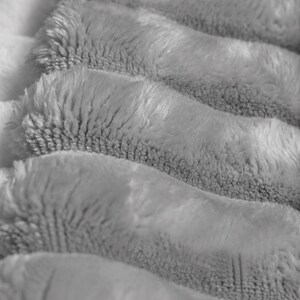 May include: A soft, light gray blanket featuring a thick, plush, and fuzzy texture. The fabric is arranged in distinct, horizontal, rounded folds that create a repeating pattern of raised ridges and deep, shadowed valleys across the surface.