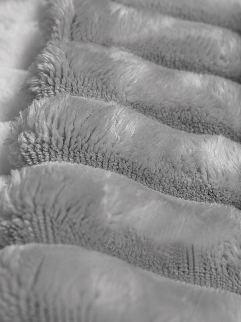 May include: A soft, light gray blanket featuring a thick, plush, and fuzzy texture. The fabric is arranged in distinct, horizontal, rounded folds that create a repeating pattern of raised ridges and deep, shadowed valleys across the surface.