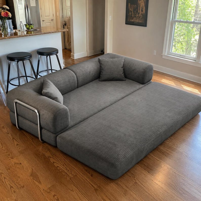 May include: A dark gray corduroy sofa bed featuring a low-profile design with rounded armrests, a metal frame support on the side, and two matching square throw pillows. The sofa is shown in an extended position, creating a flat, wide sleeping surface.