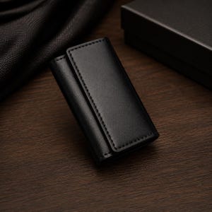 Könnte beinhalten: Ein rechteckiges Schlüsseletui aus schwarzem Leder mit glatter Oberfläche und sichtbaren, farblich abgestimmten Nähten an den Kanten. Das Accessoire verfügt über ein Klappdesign mit sicherem Verschluss und wirkt sauber und strukturiert.