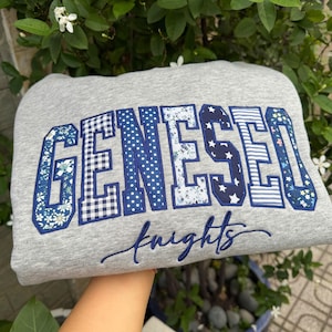 May include: A heather gray sweatshirt featuring the word GENESEO in large, block-style letters. Each letter is filled with a different blue and white patterned fabric, including floral, gingham, polka dot, and star prints. The word knights is embroidered below in blue cursive script.