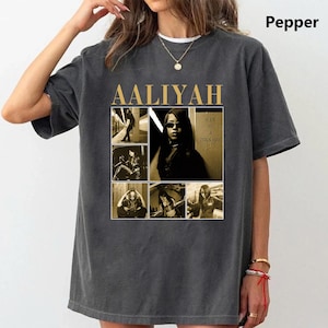 May include: Dark gray t-shirt featuring a collage of images and the name "AALIYAH" in gold lettering. The collage includes sepia-toned photos of a woman. The text "ONE IN A MILLION" is also visible.