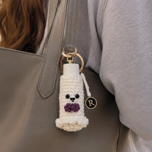 May include: A white crocheted holder for a small bottle, featuring a ghost-like face with black bead eyes, a stitched mouth, and a small purple bow tie. The holder is attached to a gold-toned metal keyring with a circular charm displaying the letter R.