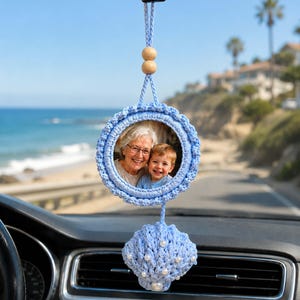 Puede incluir: Un adorno de coche tejido a ganchillo en color azul claro con un marco circular que contiene una fotografía de una persona mayor y un niño pequeño. Incluye dos cuentas de madera en el cordón y un adorno de concha tejido con perlas blancas.