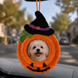 May include: A crocheted orange pumpkin car charm featuring a black witch hat with a purple band and orange buckle, a green leaf, and a black jack-o'-lantern face. The center of the pumpkin contains a circular photo of a small, light-colored dog.