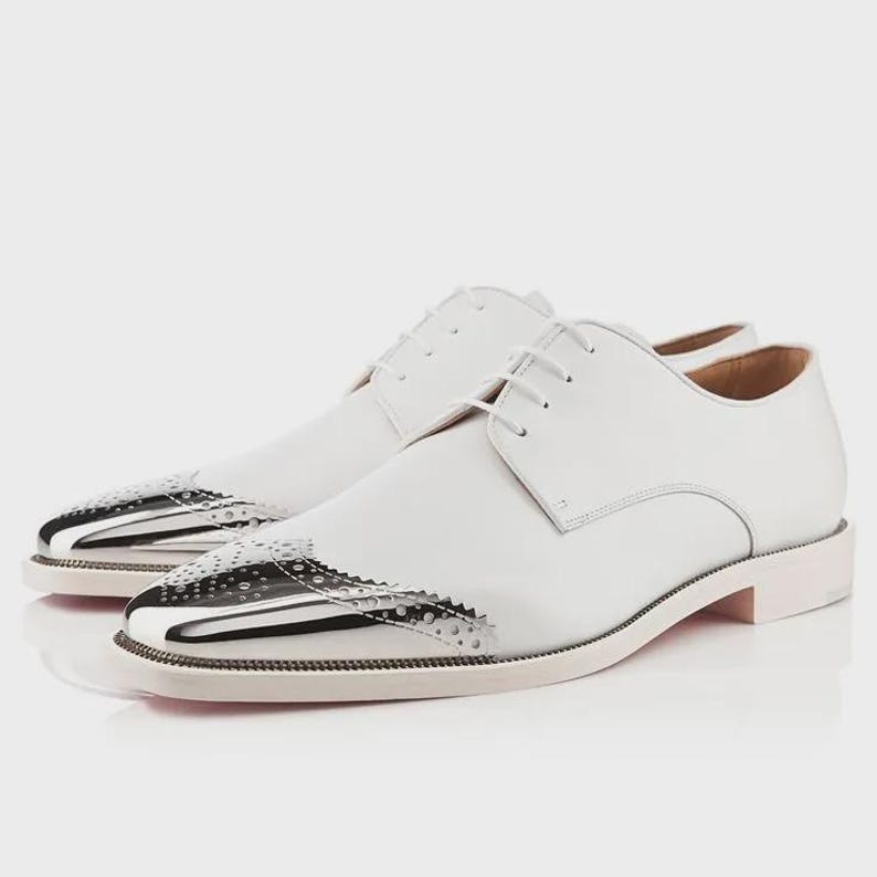 May include: A pair of white leather dress shoes with a polished silver toe cap. The shoes have a classic lace-up design and a low heel. The toe cap features a decorative perforated pattern.