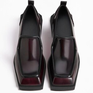 May include: A pair of dark burgundy loafers with a high-shine finish, featuring a distinct square toe, a raised apron seam, and a thick black rubber sole. The shoes have a pull tab at the heel and a smooth, structured silhouette.
