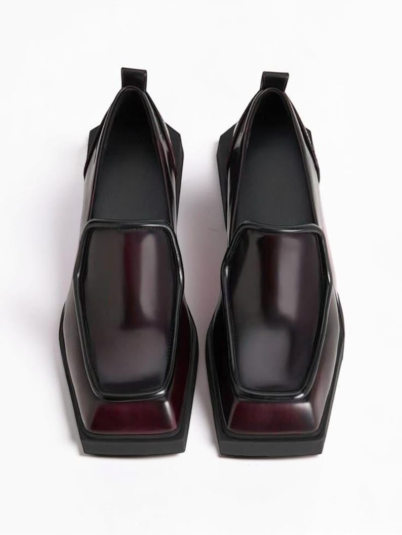 May include: A pair of dark burgundy loafers with a high-shine finish, featuring a distinct square toe, a raised apron seam, and a thick black rubber sole. The shoes have a pull tab at the heel and a smooth, structured silhouette.