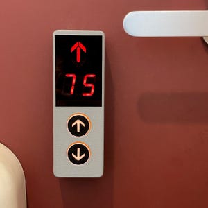 May include: A rectangular elevator control panel with a light gray frame and a black digital display showing the number 75 in red with a red upward arrow above it. Below the display are two circular buttons with white upward and downward arrows.