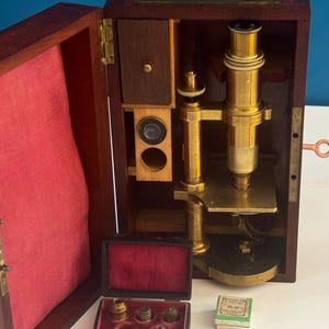 Puede incluir: Un microscopio de latón guardado en una caja de madera oscura con forro de tela roja. El conjunto incluye una pequeña caja negra con interior de terciopelo rojo que contiene cuatro lentes de latón y una pequeña caja cuadrada blanca con la etiqueta E. COUTAC 50.22 PARIS.