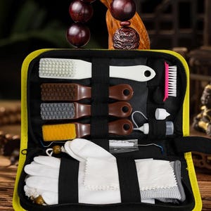 Könnte beinhalten: Eine schwarze Transporttasche mit gelbem Rand, die ein Pflegeset enthält. Das Set umfasst eine weiße Bürste, drei braune Bürsten mit weißen, grauen und gelben Borsten, eine kleine rosa Bürste, ein weißes Tuch und eine kleine Flasche.